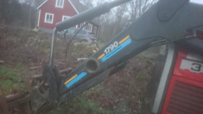 Skadad grävmaskin vid stengärde med hus och bil i bakgrunden.