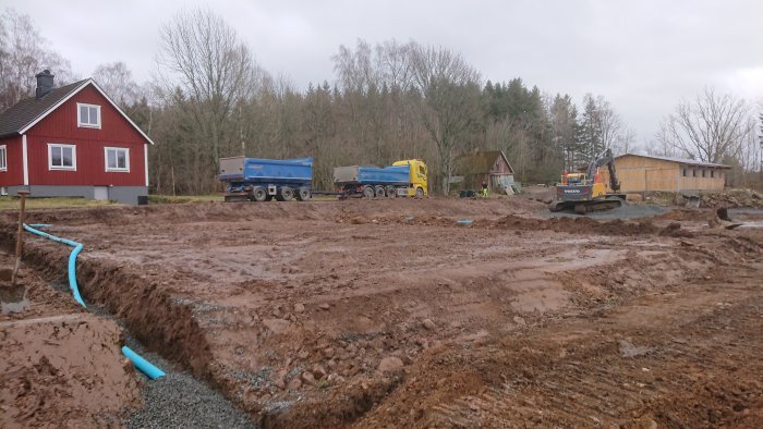 Byggarbetsplats för anläggning av paddock med grävmaskin, lastbilar och grus, med röd stuga i bakgrunden.