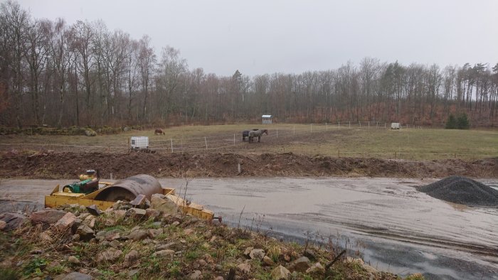 Byggnation av paddock, grävmaskin bredvid väg, högar med makadam, hästar på fält i bakgrunden.