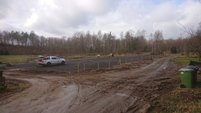 Pågående arbete med paddockbygge, visar markerade stolpplatser, utspridd makadam, en grävmaskin och en pickup.