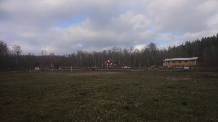 Vy över pågående paddockbygge med rödfärgat hus och stall i bakgrunden samt maskiner och material på fält.