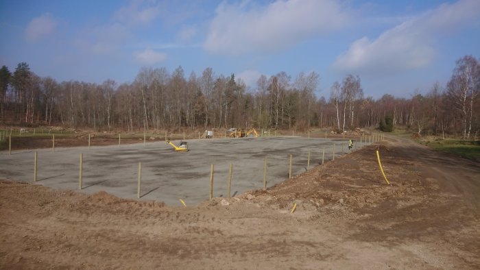 Ett nytt byggprojekt: färdigpaddad stenyta med utslagna stolpar och nedgrävda kabelrör till höger, omgiven av skog.