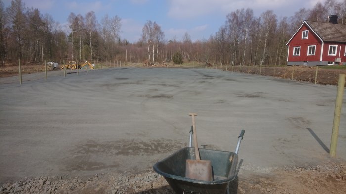 Nyplanerad och paddad mark med stolpar och kabelrör, skottkärra i förgrunden, röd stuga i bakgrunden.