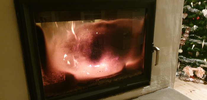 Nyligen rengjort kaminfönster med synliga flammor och lätt sotning, intill en julgran.