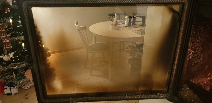 En inomhusbild med blick på en rund träbord med stolar genom en suddig glasruta, nära ett juldekorerat träd.
