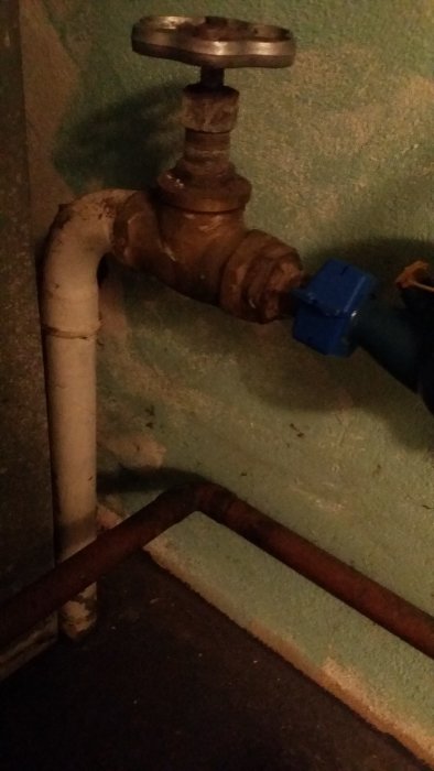 VVS-installation med vita och rostiga rör samt en blå ventil i ett mörkt utrymme.