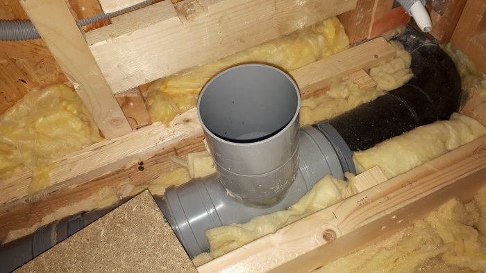 Installationsbild på ett grått avloppsrör med T-förgrening under bjälklag isolerat med gul mineralull.