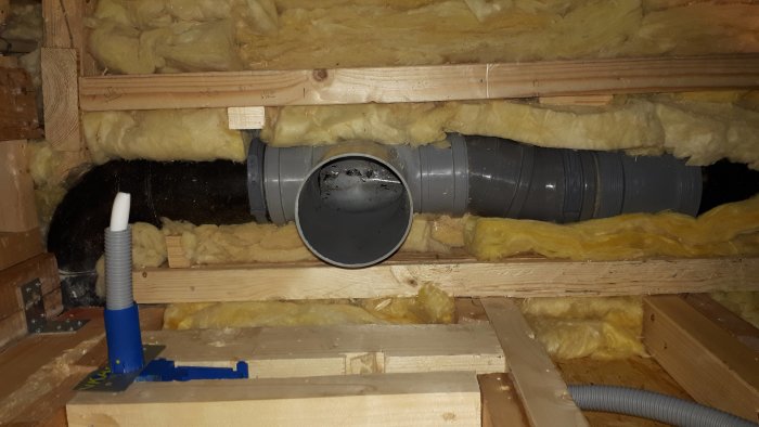 Ventilationsrör monterat mellan träbjälkar, omgivet av gul isolering i en byggnadskonstruktion.