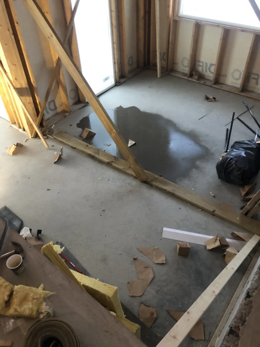 Vattenpöl på golv i ofärdigt byggprojekt med synliga trästommar och skräp.