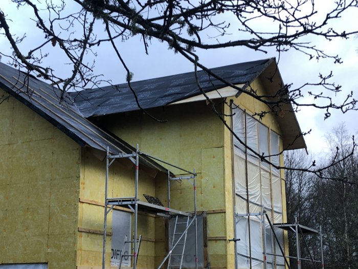 Halvfärdigt hus med byggnadsställningar och oskyddad takkonstruktion under mulet väder.