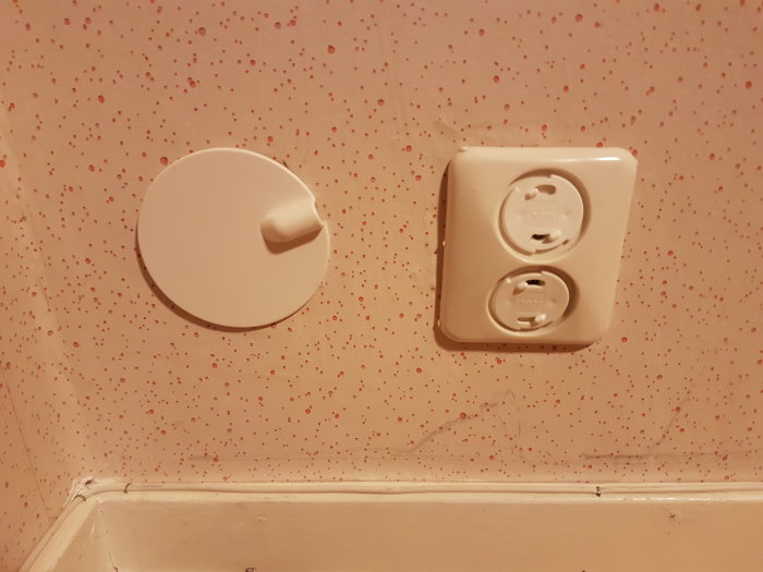 Vägg med präglad tapet, eluttag och en rund, vit lucka mot hörnet av rummet.