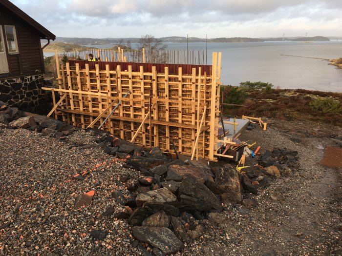 Byggställningar och träformar för gjutning av betongväggar på en tomt med havsutsikt och klippor i förgrunden.