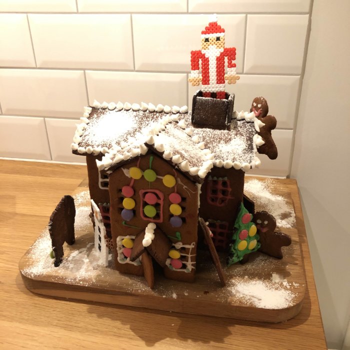 En handgjord pepparkakshusreplika med färgglada dekorationer, glasyr, en pepparkaksgubbe framför, på ett träunderlag.