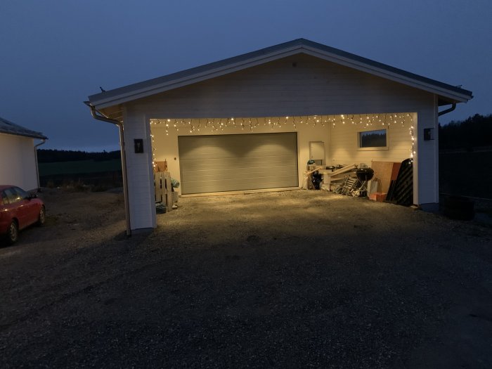 Exteriör av vit garagebyggnad i skymningen med tända ljusslingor och parkerad bil.