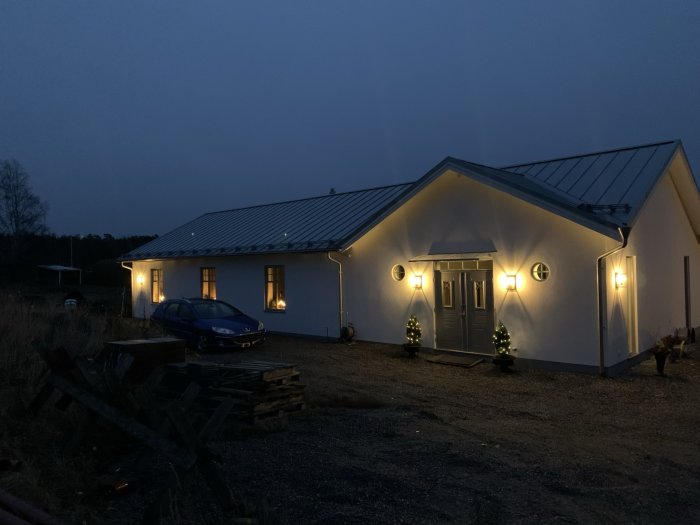 Exteriör kvällsbild av ett modernt hus med belysning, carport och juldekorationer vid entrén.