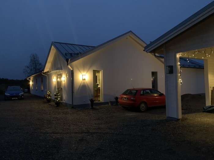 En modern bostad på kvällen med julbelysning, carport och en röd bil parkerad utanför.