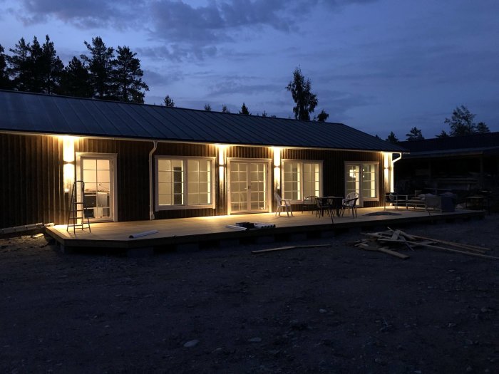 Nybyggt hus av lösvirke under skymningen med tänd utomhusbelysning och vidbyggt garage och carport.