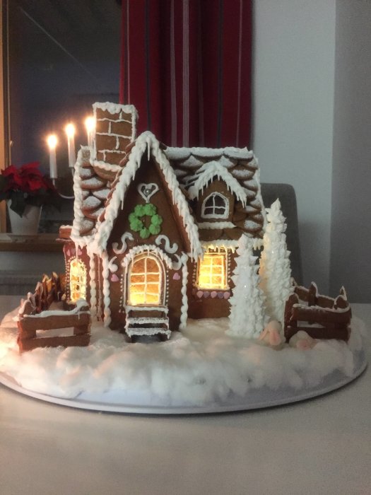 Hemgjort pepparkakshus med ljus inuti, snöeffekt och juldekorationer mot en fönsterbakgrund.