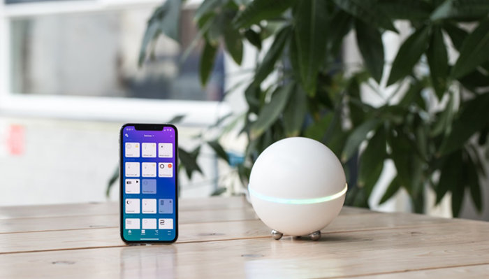 Smartphone med öppen app bredvid en sfärisk Homey smart home hub på ett träbord mot en bakgrund med växter.