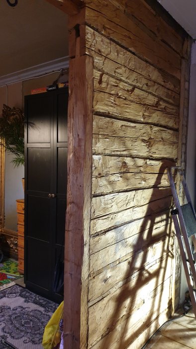 Rustikt träväggsparti med synliga stockar och en stege vid sidan, inne i ett hus.
