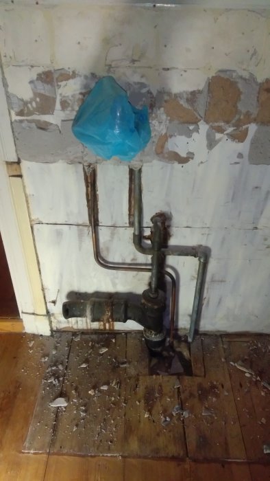 Gamla vattenrör och ventiler mot sliten vägg, täckt med blå plastpåse, renoveringsarbete pågår.