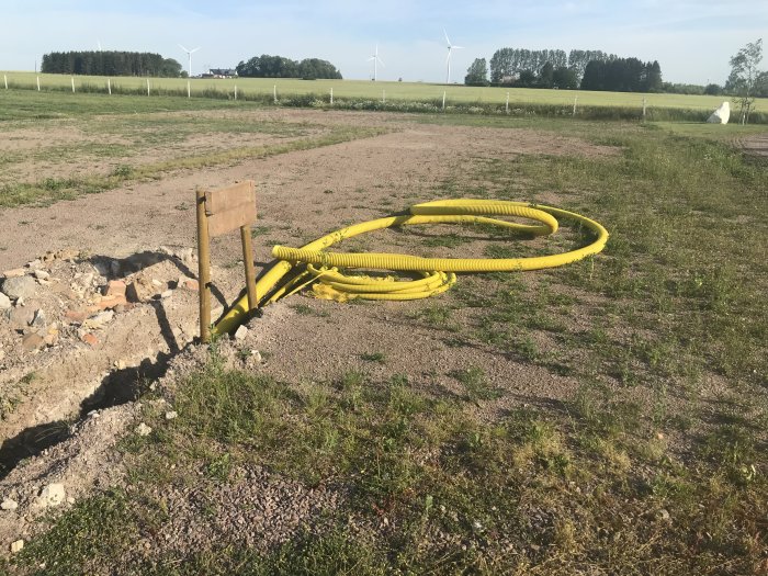 En öppen byggarbetsplats med gula elkablar och ett avtaget staket, framför en lantlig bakgrund.