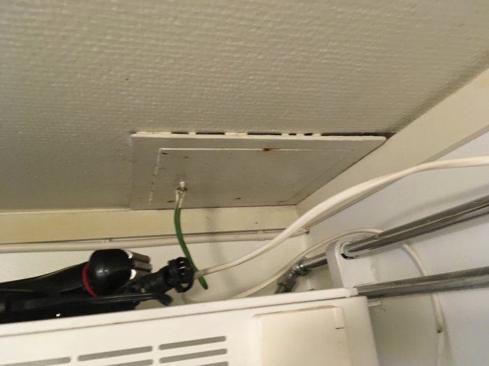 Ventilationsutgång i taket ovanför en lägenhetsdörr, kantad av kablar och en fläkt vid sidan.