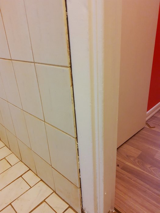 Kaklad vägg med springor vid möte med en vit dörrkarm och ovanför en beige kakelrad.
