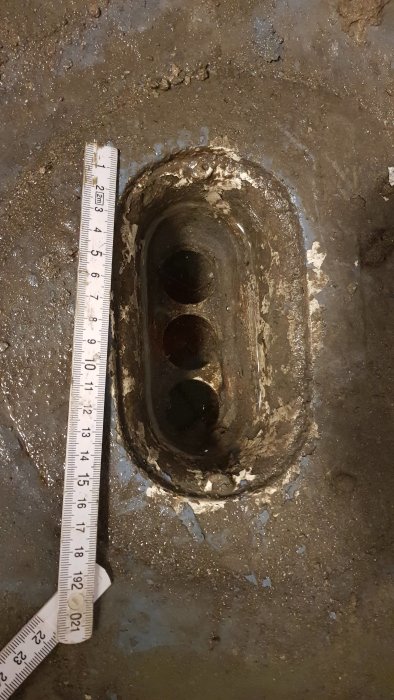 Täppt golvbrunn i betong med synliga avlagringar och en mätsticka som visar djupet.