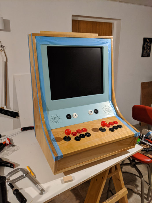 Hembyggt arkadspel med lackerad plywood och körsbärspaneler, joystick och knappar, i ett arbetsrum.