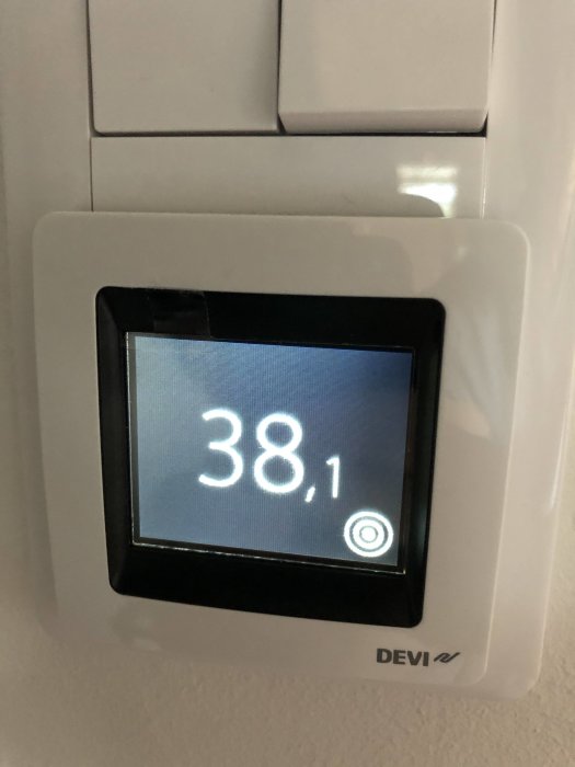 Termostat visar 38,1 grader, troligtvis för golvvärme, på en vit vägg.