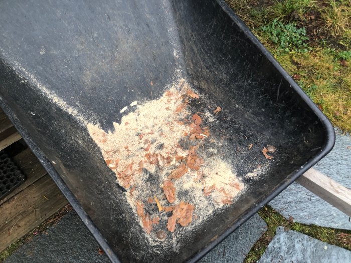 Svart skottkärra med vedspån och några små vedbitar, utomhus på trädäck och gräs i bakgrunden.