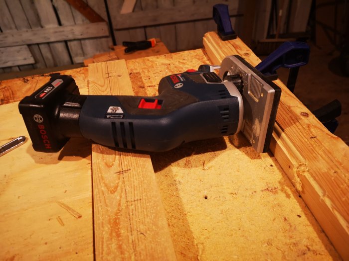 En handhållen Bosch-fräs ligger på ett arbetsbord bredvid nyligen frästa träbrädor med spån och verktyg.