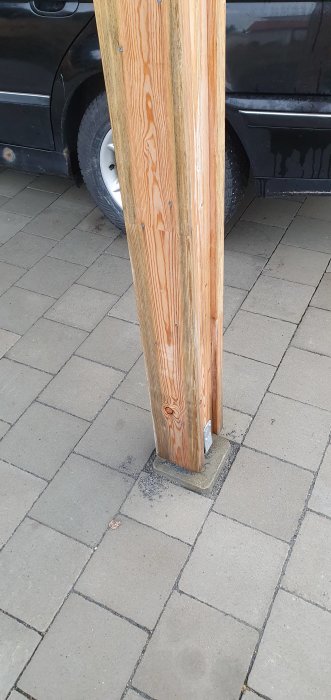 Trästolpe till en carport med sporadisk grön växtlighet vid basen, stående på en stenlagd uppfart med en bil i bakgrunden.