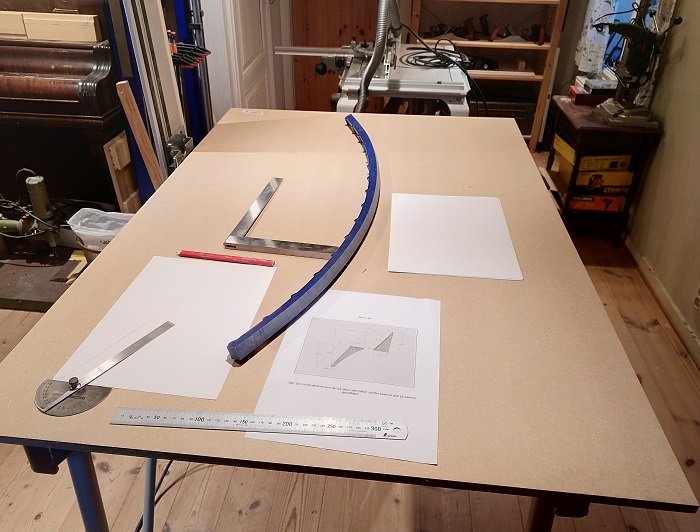 Arbetsbänk med ritningar, linjal, fräsmall och blyertspenna i ett snickarrum.