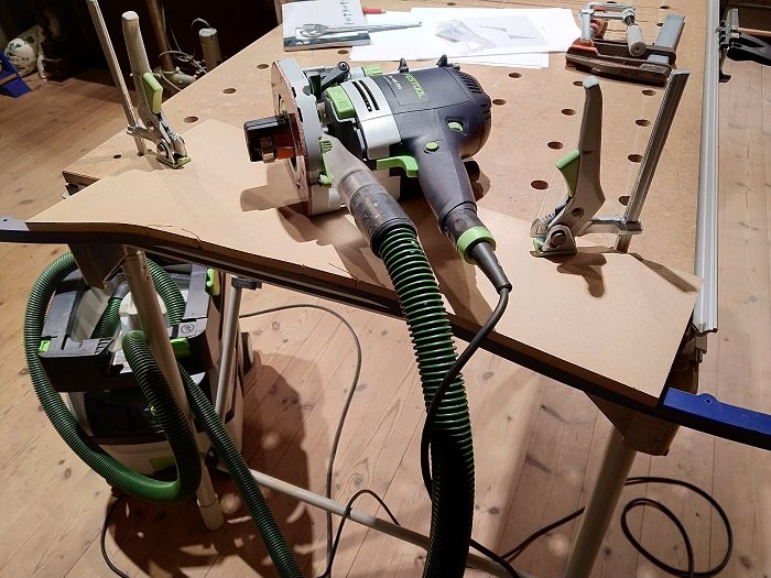 En överfräs fastspänd på en arbetsbänk med utfräst mall i trä och kopplad till en dammsugarslang.
