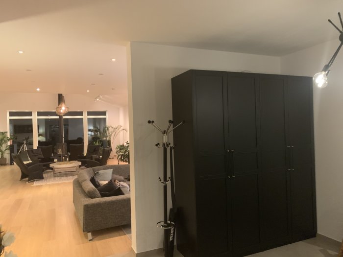 Vy över hall med svart garderob och blick in i ett vardagsrum med soffa och fåtöljer.