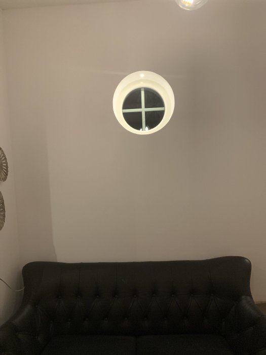 Rund väggfönster ovanför en mörk tuftad soffa i en hall med neutral väggfärg.
