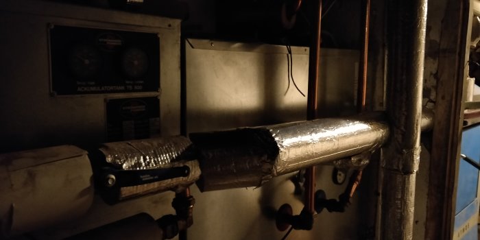 Isolerade rör och ackumulatortank i ett mörkt teknikutrymme där tanken står lägre än rören.