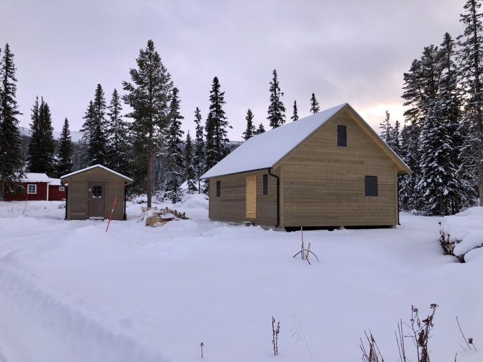 Nybyggt hus och förråd i snötäckt landskap med skog i bakgrunden under gryning eller skymning.