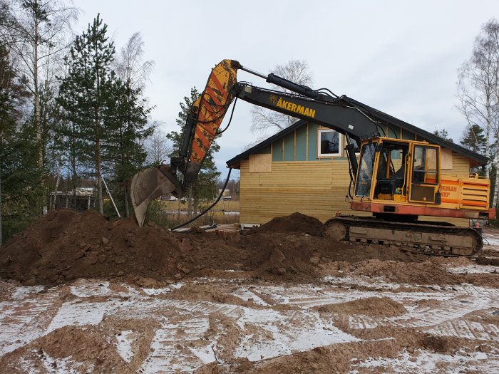 Grävmaskin som gräver ner dagvattenledningar vid ett hus med delvis snötäckt mark.