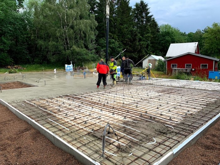 Byggarbetsplats med gjutet mellanbjälklag och armeringsjärn, arbetare, betongbil och villavagn i bakgrunden.