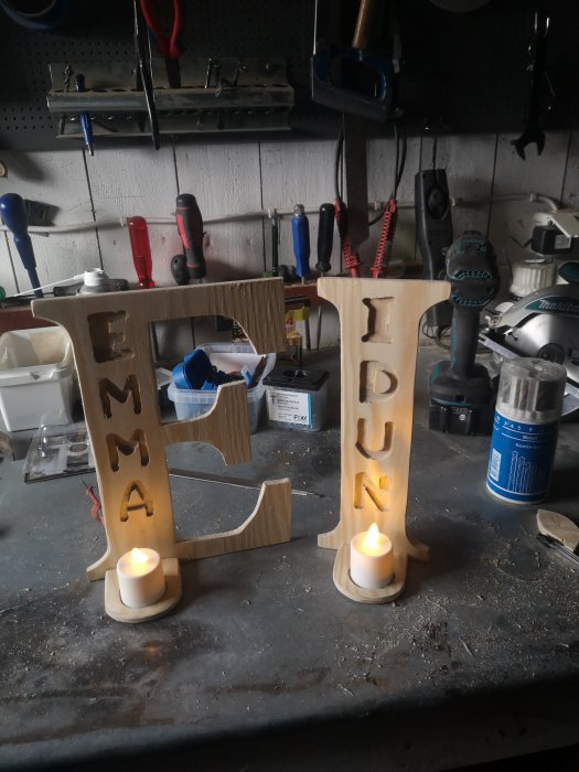 Handgjorda trädekorationer med namnen "EMMA" och "IDUN" i en verkstad, med tända ljus.