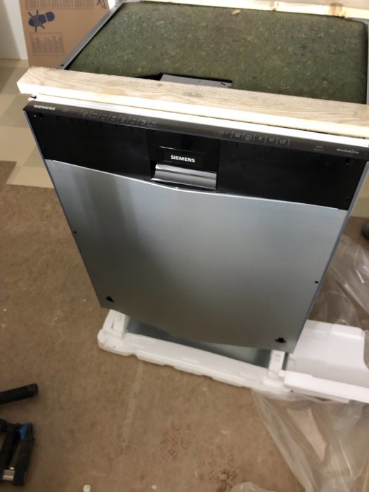 Damaged Siemens dishwasher with dented front and door standing on a floor with packaging materials around.