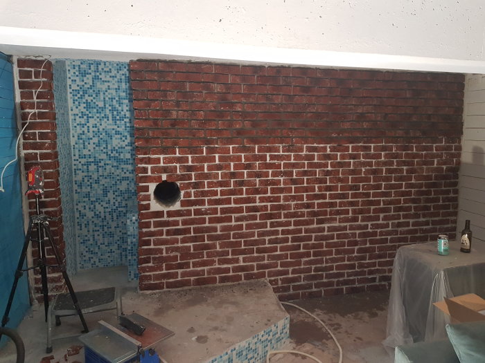 En pågående renovering av ett rum med en delvis plattsatt vägg som liknar tegel och en delvis täckt vägg med kakelmosaik.