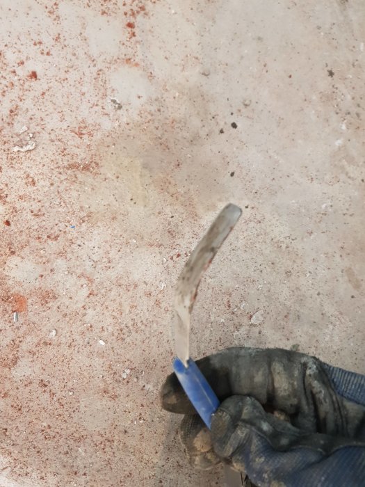 Hand i arbets handske håller en modifierad, vinklad fogkniv över betonggolv.