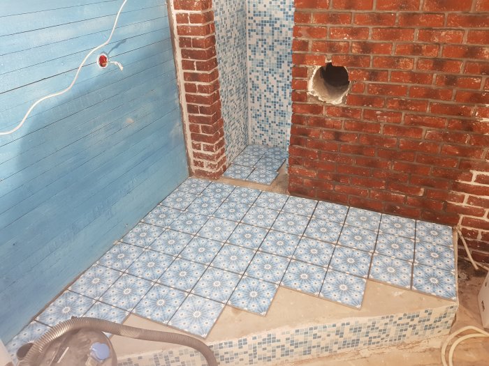 Renoveringsprojekt med blå-vita kakelplattor på golv och mosaikplattor på podie vid tegelvägg.