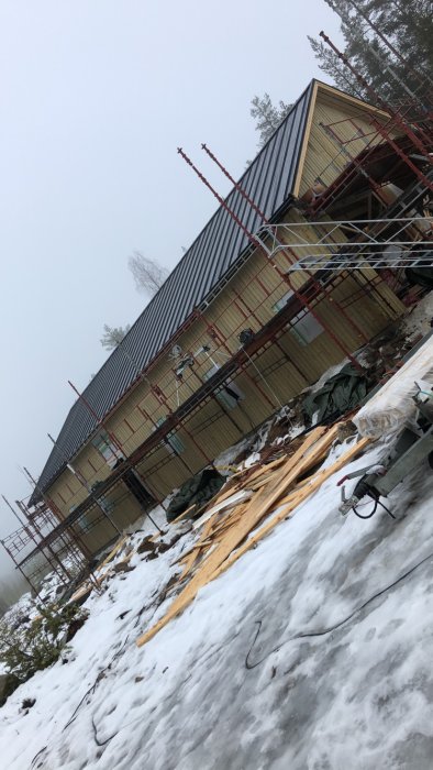 En sned bild av ett hus under konstruktion med ställningar och snö på marken.