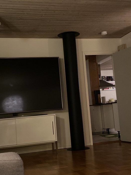 Ett svart skorstenrör i ett vardagsrum som sträcker sig upp mot ett träpaneltak, intill en vit TV-bänk.