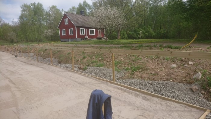 Nybyggd paddock med pågående konstruktion av staket, makadamgrund och en upphängd skjorta i förgrunden, lantligt röd hus i bakgrunden.